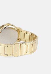 Orologio da polso in tono oro con un elegante cinturino metallico, caratterizzato da anelli rettangolari e una cassa lucida. Il retro è liscio e riflettente, mostrando il marchio.