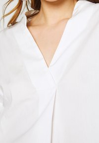 Bílá halenka s límečkem a výstřihem do V, s minimalistickým designem a hladkým, lehkým materiálem. Viditelné švy jako akcent.