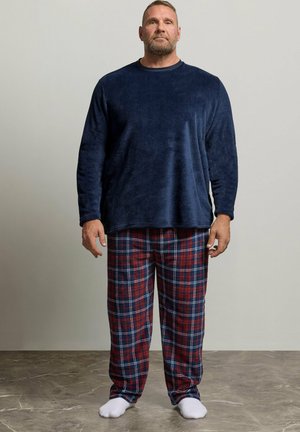 Homme avec barbe portant un haut à manches longues bleu marine et un pantalon de pyjama à carreaux rouges et bleus, debout pieds nus sur un sol gris.