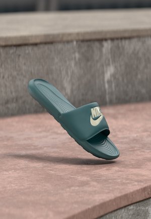 Tumeroheline Nike slaid-sandaal tekstuurse tallaga ja suure valge Nike logoga rihmal, kujutatud kivipinna kohal hõljuvana.