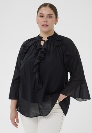 Kobieta ubrana w przezroczystą czarną bluzkę z falbankami i rękawami w kształcie dzwonu, w zestawieniu z białymi spodniami ozdobionymi metalowymi guzikami.