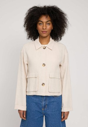 Woman with curly hair wearing a beige buttoned jacket with front pockets and blue jeans, standing against a plain background.