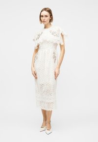 Γυναίκα που στέκεται φορώντας λευκό midi φόρεμα από δαντέλα με ώμους κάπα και λευκές μυτερές γόβες με λουράκια στον αστράγαλο.