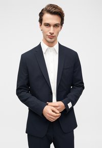 Μπλε σκούρο σακάκι με δομημένους ώμους, γιακάδες με περικοπή και κλείσιμο με ένα κουμπί, συνδυασμένο με ένα λευκό πουκάμισο. Ορατή λεπτομέρεια με τσέπη στο εμπρόσθιο μέρος.