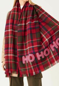 Écharpe en tartan rouge et vert avec des bords frangés, ornée de lettres roses "HO HO HO". Fabriquée en tissu doux, de grande forme rectangulaire.