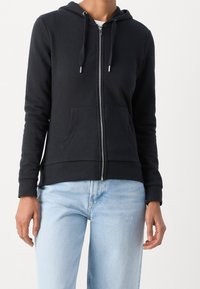 Schwarze Zip-Hoodie aus weichem Baumwollmaterial mit einer Fronttasche, Kordelzugkapuze und gerippten Bündchen. Kombiniert mit hellblauen Jeans.