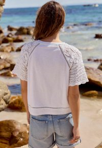 Weißes T-Shirt mit Spitzenrüschenärmeln und strukturiertem Saum, kombiniert mit hellblauen Jeansshorts, vor einem Strandhintergrund.