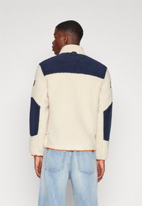 Fleece jacket in cream and navy, featuring textured fabric, high collar, and orange stitching along the hem. Distinctive color blocks on sleeves.