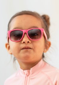 Vaaleanpunaiset aurinkolasit tummilla linsseillä; muoviset kehykset. Lapsi, jolla on päällään vaaleanpunainen vetoketjullinen paita. Läheltä otettu, neutraali ilme, yksinkertainen tausta.