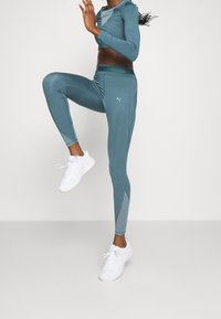 Kvinna i teal träningstights och crop top som höjer ett knä, iklädd vita löparskor, mot en ljus, neutral bakgrund.