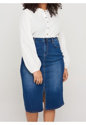 Frau trägt eine weiße Langarmbluse mit Knöpfen, die in einen hoch taillierten blauen Jeansrock mit Frontschlitz gesteckt ist.