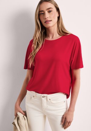 Kvinde iført en knald rød kortærmet skjorte og hvide bukser, der holder en beige jakke i den ene hånd mod en ensfarvet baggrund.