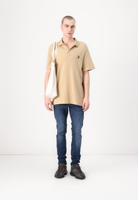 Tan polo skjorte med en liten logo, blå jeans og svarte sneakers. Modellen holder en hvit kanvas totebag. Minimalistisk hvit bakgrunn.