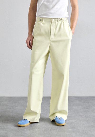 Pantaloni a gamba larga di un giallo chiaro, realizzati in cotone con una texture liscia. Presentano una vita con bottoni e tasche laterali. Abbinati a scarpe blu.