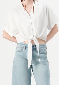 Donna che indossa una camicia bianca a maniche corte con bottoni annodata in vita e jeans a vita alta blu chiaro su uno sfondo bianco.