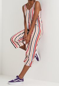 Combinaison rayée à coupe loose, présentant des lignes verticales en rouge, orange, blanc et noir. Associée à des baskets violettes.