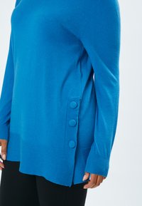 Blauer Strickpullover mit langen Ärmeln, seitlichen Knopfdetails und geripptem Saum. Weicher Stoff und lockere Passform, ausgestattet mit drei großen Knöpfen.