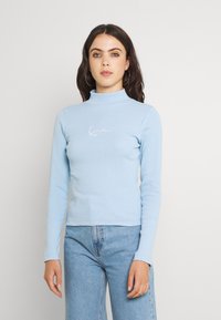 Maglione a collo alto a maniche lunghe di un blu chiaro con texture a coste, caratterizzato da un logo ricamato bianco sul petto, abbinato a jeans blu.