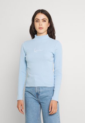 Svijetlo plavi dugi rukav turtleneck top u rebrastoj teksturi, s bijelim vezom brenda na prsima, u kombinaciji s plavim trapericama.