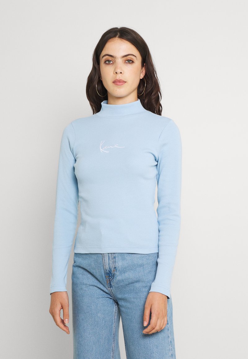 Maglione a collo alto a maniche lunghe di un blu chiaro con texture a coste, caratterizzato da un logo ricamato bianco sul petto, abbinato a jeans blu.