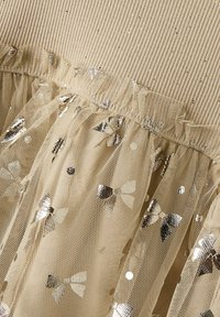 Beige Rippenoberteil mit einem Rüschentüllüberzug, der silberne Pailletten-Bowmuster aufweist und eine weiche, strukturierte Optik hat.