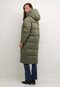 Olīvu zaļš pūkains mētelis ar kapuci, garš siluets, quilted tekstūra un pastiprināta šuve. Apvienots ar tumšām džinsu biksēm un augstiem zābakiem.
