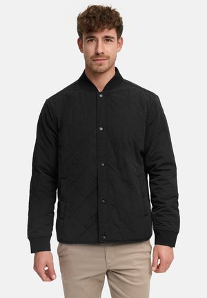 Uomo con capelli corti e ricci che indossa una giacca nera trapuntata con bottoni e pantaloni beige su uno sfondo semplice.