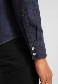 Tmavě modrá džínová košile s dlouhými rukávy, vystuženými stříbrnými patentkami na manžetách, viditelným šitím a střihovaným střihem.