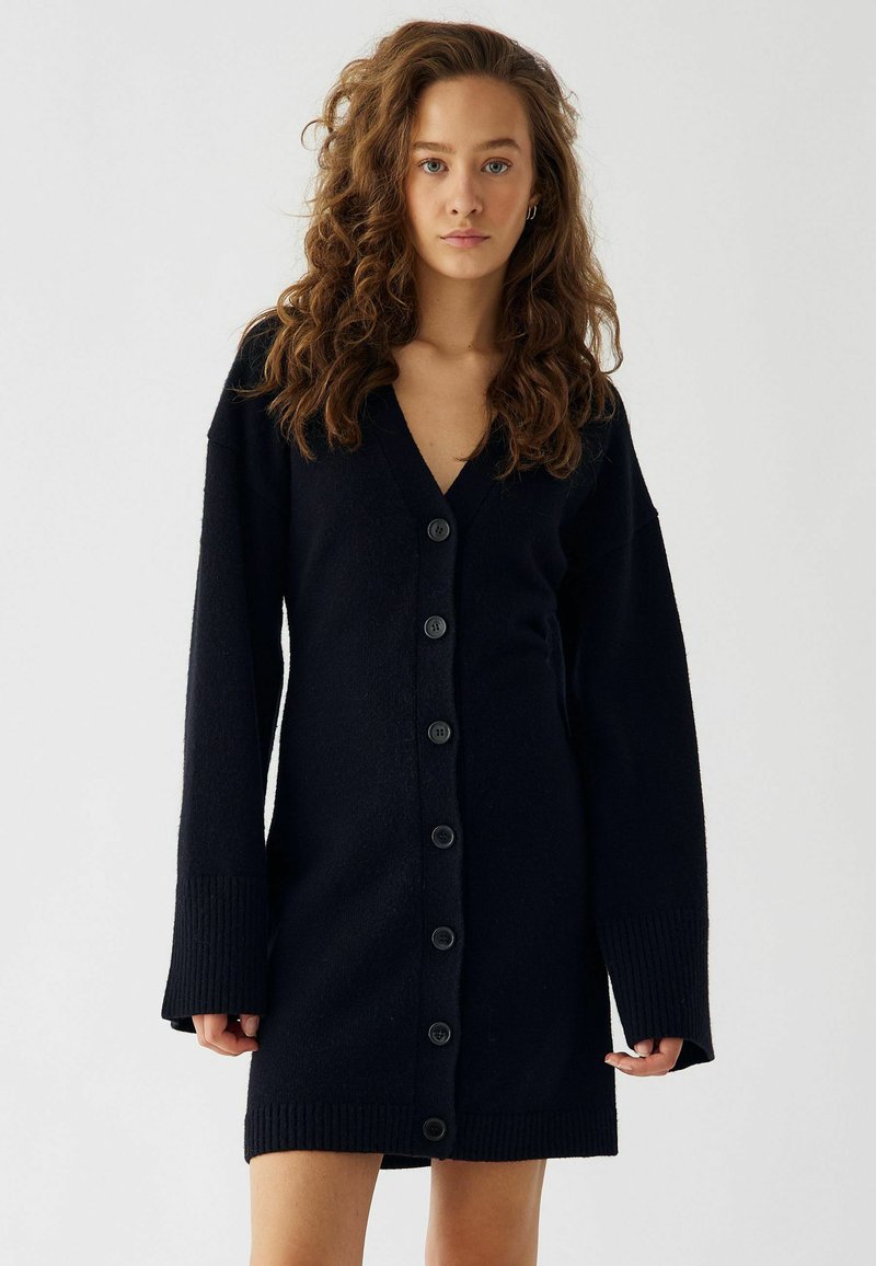 Melnas, garās piedurknes kardigāna kleita ar V-veida kakla izgriezumu, kas ir ar piecām lielām pogām priekšpusē un ribota apdare uz piedurknēm un apakšmalas.