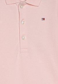 Jasnoróżowa koszula polo z trzema białymi guzikami i małym haftowanym logo w czerwono-białoniebieskich kolorach po lewej stronie klatki piersiowej.