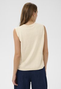 Kreminė be rankovių palaidinė su apvaliu iškirpimu, minkšta tekstūra ir raukšlėta apatine dalimi. Dizainas pasižymi minimalistiniu stiliumi, tinkamu kasdieniam dėvėjimui.