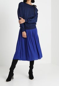 Merihenkinen kimalteleva villapaita pitkillä hihoilla, yhdistettynä laskostettuun kuninkaansiniseen hameeseen ja mustiin reisikorkeisiin saappaisiin. Yksinkertainen muotoilu ja tekstiilissä on tekstuuria.