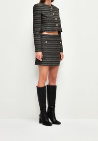 Veste cropped noire et or avec des épaules structurées, assortie d'une jupe et de bottes en cuir noir jusqu'aux genoux.