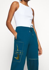 Pantalones de chándal en color verde azulado con un logo dorado de "just do it", cinturilla elástica y bolsillos laterales. Acompañados de una camiseta sin mangas blanca de canalé.