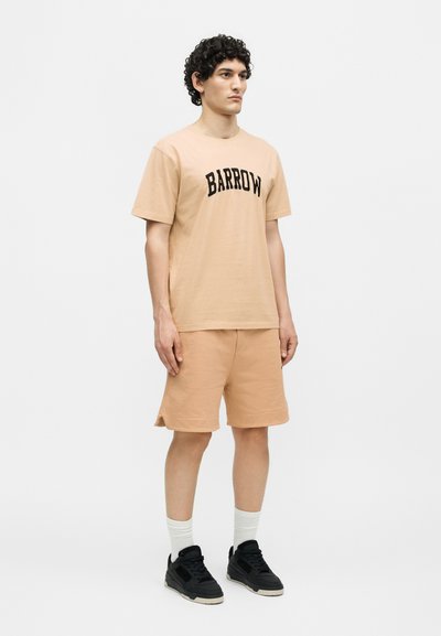 Νέος άνδρας με μπεζ σορτς και ταιριαστό μπλουζάκι "BARROW" με λευκές κάλτσες και μαύρα αθλητικά παπούτσια, στέκεται μπροστά σε απλό λευκό φόντο.