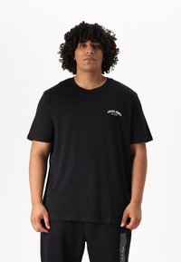 Młody mężczyzna o kręconych włosach, ubrany w czarny t-shirt Jack & Jones oraz czarne spodnie Calvin Klein, na jednolitym białym tle.