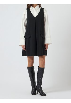 Femme portant une robe noire sans manches à col en V sur une chemise blanche à manches longues, associée à des bottes noires à talons montant jusqu'aux genoux, debout devant un fond blanc.