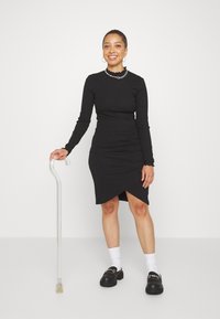 Svart långärmad klänning med rynkade detaljer och asymmetrisk nederkant. Kombinerad med svarta platåskor och en klar käpp.