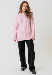 Gaiši rozā adīta džemperis ar kabelīšu rakstu, pārmērīgs piegriezums, ribotām piedurknēm un apakšmalu. Saskan ar melnām platām biksēm un spīdīgām kurpēm.