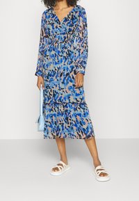 Maxi mekko, jossa on pitkät hihat, sininen ja vaaleanpunainen abstrakti kuosi, V-päänti, laskostettu vyötärö ja kerroksellinen helma. Yhdistetty valkoisten sandaalien ja sinisen käsilaukun kanssa.