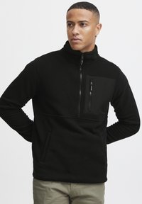 Svart fleece-pullover med høy krage, glidelås foran og brystlomme. Har sidelommer og strukturert stoff. Moderne passform.