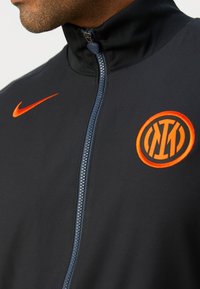 Gros plan d'une personne portant une veste noire zippée avec un logo Nike orange et le logo du club de football de l’Inter Milan sur la poitrine.