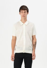 Pleteni kratkotrajni majica v off-white barvi, ki vsebuje vzorec fine mreže in diamantnih oblik, s klasičnim ovratnikom in zapiranjem na gumb.