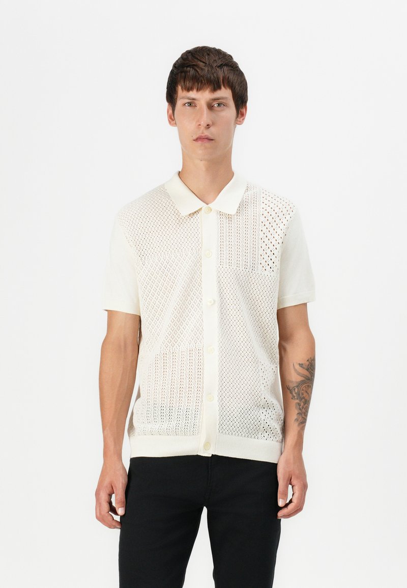 Pleteni kratkotrajni majica v off-white barvi, ki vsebuje vzorec fine mreže in diamantnih oblik, s klasičnim ovratnikom in zapiranjem na gumb.