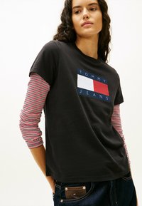 Czarna koszulka z niebieskim, czerwonym i białym logo "TOMMY JEANS", nałożona na czerwoną i białą koszulę z długim rękawem. Widać dżinsowe spodnie.