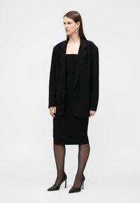 Γυναίκα που φορά μαύρο φαρδύ σακάκι πάνω από μαύρο φόρεμα χωρίς τιράντες, μαύρες διαφανείς καλσόν, μαύρες ψηλοτάκουνες γόβες και μακριά ασημένια σκουλαρίκια, όρθια.