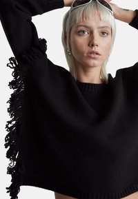 Černý zkrácený svetr s texturovanými třásněmi podél spodního lemu a rukávů. Má kulatý výstřih a ribbované okraje.