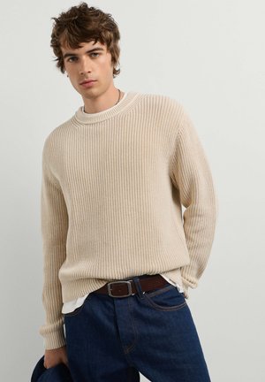 Junger Mann mit welligem braunem Haar, der einen beigen gestrickten Pullover, dunkelblaue Jeans und einen braunen Gürtel trägt und eine dunkelblaue Jacke hält.