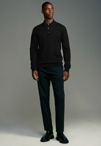 Melnā džemperis ar apkakles izgriezumu un trim pogām, apvienots ar tumši zaļām biksēm un melniem potītes zābakiem. Gluda tekstūra.