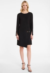 Robe noire en tissu doux avec un col rond, des manches longues à poignets fendus, et un ourlet diagonal accentué par un détail en anneau doré.